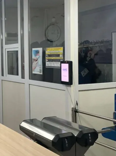 A slim wall tablet showing 'AMS' on a pink screen, mounted on a glass partition beside a checkpoint turnstile