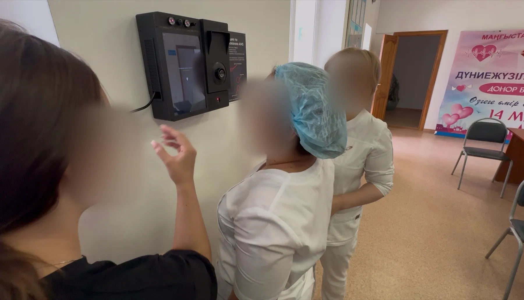 Two workers in white uniforms, one in a hair cover, facing the AMS wall unit at a check station while another person points at its screen; faces are blurred