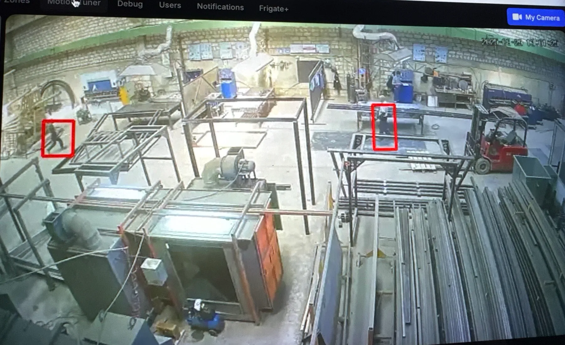 A camera view of a metal fabrication workshop in the Frigate interface, two people outlined in red boxes among steel frames and a forklift
