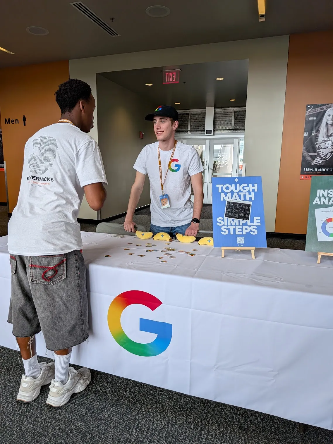 Two people talking across a table draped with a large Google 'G', one of them in a lanyard and a Google T-shirt