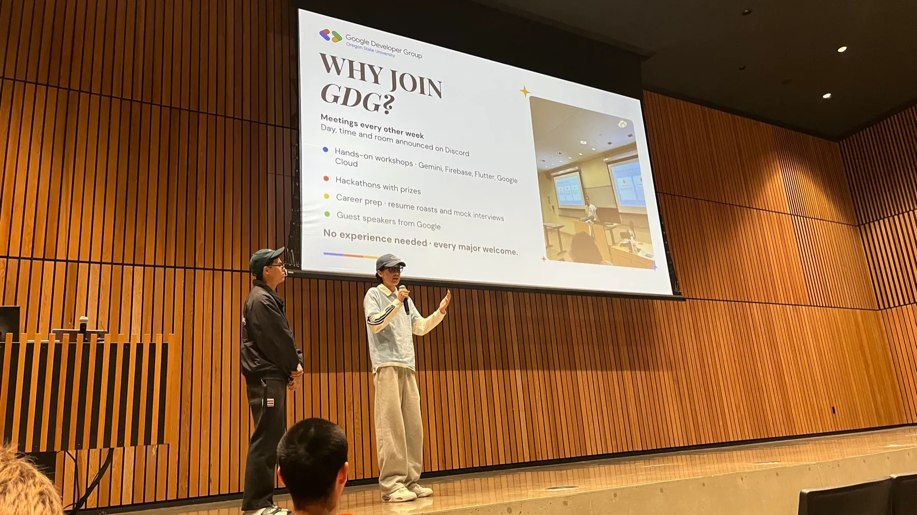 Jak holding a microphone on a wood-panelled stage, a second person standing behind, under a projected slide headed 'Why join GDG?'
