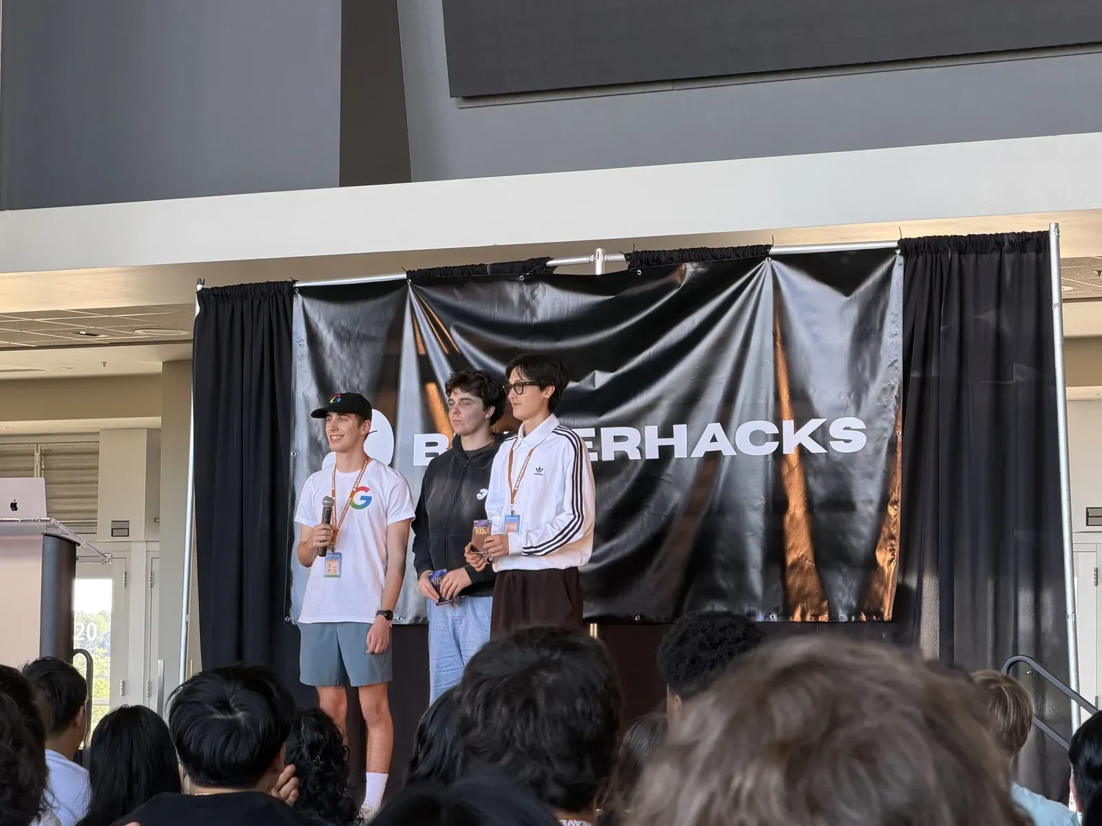 Jakhangir and his teammate on stage under the BeaverHacks banner, both holding prize cards, beside a presenter with a microphone, the audience in the foreground