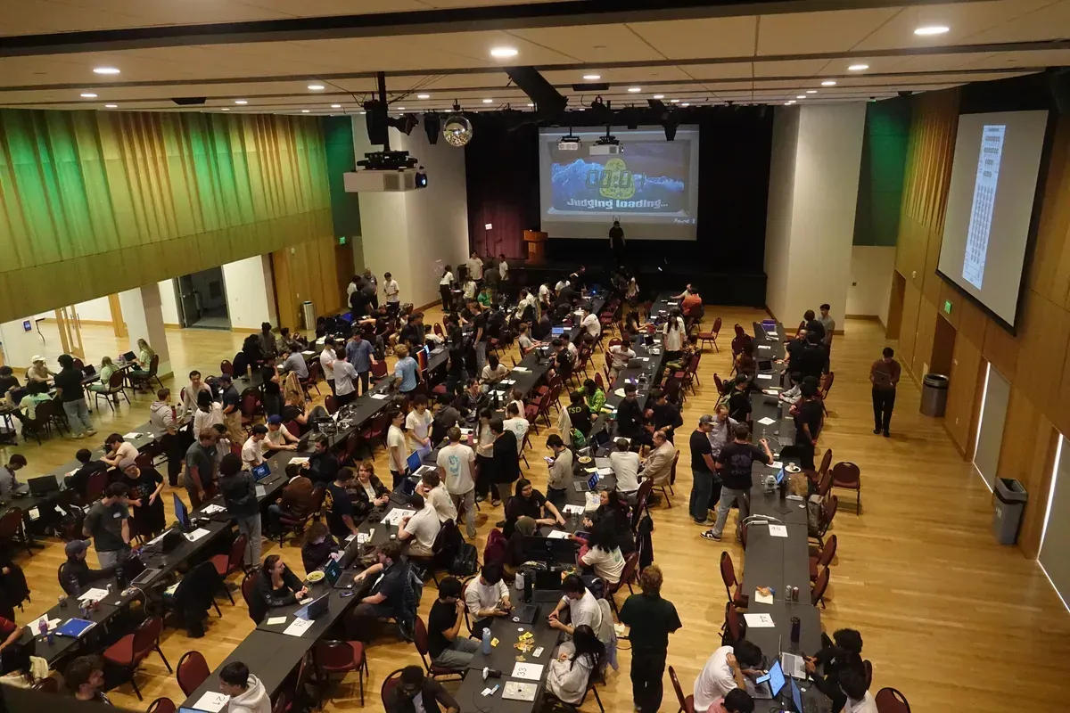 The QuackHacks 3 hall from above: rows of tables full of hackers, the stage screen at the far end